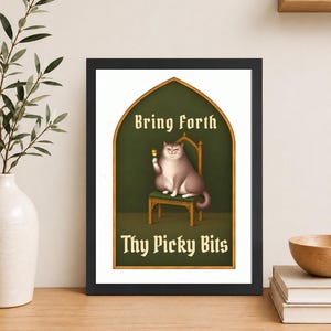 May include: A framed wall art print featuring a grey cat sitting on a wooden chair inside a gold-arched frame. The cat holds a small skewer. The text reads Bring Forth Thy Picky Bits in a gothic font against a dark green background.