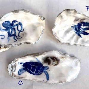 Könnte beinhalten: Drei Austernschalen mit silberfarben bemalten Rändern. Schale A zeigt eine blaue Krakenillustration. Schale B zeigt ein blaues Meeresschildkrötenmotiv. Schale C zeigt eine blaue Meeresschildkrötengrafik. Jede Schale ist mit einem blauen Buchstaben markiert.