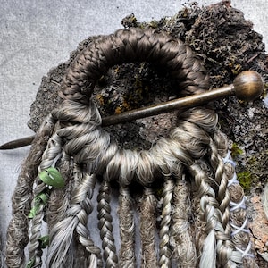 Könnte beinhalten: Ein Haaraccessoire mit einem kreisförmigen Ring, der mit braunen Fasern umwickelt und mit einer dunklen Holznadel befestigt ist. Daran hängen mehrere Strähnen aus braunem und grauem Kunsthaar, darunter Zöpfe, gedrehte Strähnen und lockige Partien.