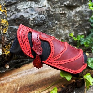 Könnte beinhalten: Ein zylindrisches Etui aus schwarzem Leder mit einem Überzug aus dunkelroten Lederpaneelen, aufwendigen Ausschnitten und dekorativen roten Nähten. Ein kleiner roter Lederriemen mit einem blattförmigen Anhänger ist seitlich befestigt.