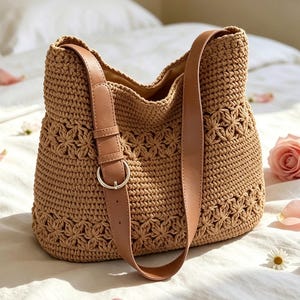 May include: A tan crochet shoulder bag featuring a textured floral pattern throughout the body. The bag includes a smooth tan faux leather strap with an adjustable silver-toned metal buckle, providing a structured look to the soft woven material.
