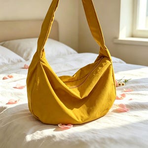 Puede incluir: Un bolso hobo de color amarillo mostaza con una correa ancha para el hombro y cierre de cremallera superior. El bolso presenta una estructura suave y holgada con una silueta de fondo curvo y un exterior de tela lisa de color sólido.
