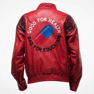 May include: A red leather jacket featuring a large embroidered patch on the back depicting a blue and red pill. The text GOOD FOR HEALTH BAD FOR EDUCATION is embroidered in a circular pattern around the pill graphic. The jacket has dark red shoulder panels.