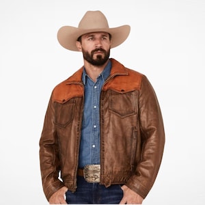 May include: A beige felt cowboy hat paired with a brown leather jacket featuring a contrasting orange-brown suede yoke across the shoulders and chest. The jacket includes a front zipper, two chest pockets with flaps, and a blue button-down shirt underneath.
