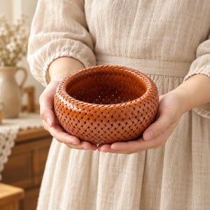 May include: A small, round, woven basket with a warm, reddish-brown finish. The basket features a tight, intricate diagonal lattice pattern throughout its exterior, creating a textured surface with a smooth, polished appearance and a wide opening.