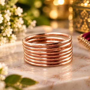 May include: A stack of seven thin, circular copper-colored bangles with a smooth, polished finish. The bracelets are arranged in a vertical column, showcasing a consistent metallic sheen and rounded profile across each individual band in the set.