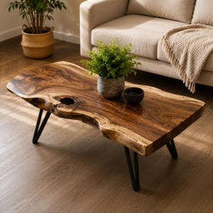 Peut inclure: Une table basse à bord brut présentant une surface en bois naturel avec une finition brun foncé et un nœud central. La table est soutenue par quatre pieds en épingle en métal noir. Une petite plante verte et un bol noir sont posés dessus.