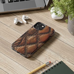 May include: A smartphone case featuring a brown, quilted leather-like texture with a diamond-stitched pattern. The case has a precise cutout for the dual-lens camera system on the back, maintaining a sleek profile while protecting the device.