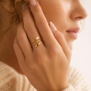 May include: A gold-toned bypass ring featuring a sleek, multi-layered design with tapered ends that wrap around the finger. The polished metal surface reflects light, creating a smooth, continuous appearance across the three visible bands.