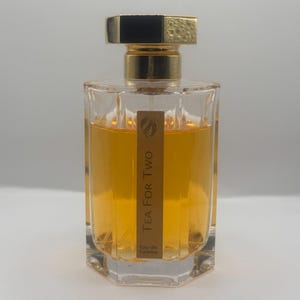 May include: A rectangular glass perfume bottle filled with amber-colored liquid. The bottle features a vertical gold label with the text Tea For Two Eau de Toilette and a gold cap with a black top. The glass has a faceted, multi-sided design.