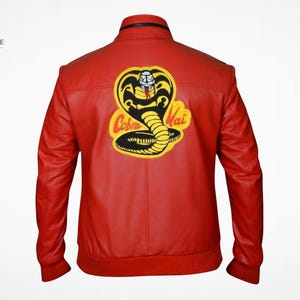 Puede incluir: Una chaqueta de cuero roja con un gran parche bordado en la espalda. El parche muestra un gráfico de una cobra negra y amarilla con el texto Cobra Kai en letras rojas. La chaqueta tiene cuello alto, mangas largas y puños acanalados.