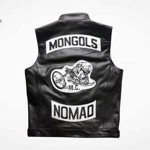 May include: A black leather vest featuring white patches on the back. The top patch reads MONGOLS, the center patch depicts a cartoon character riding a motorcycle with the text M.C. below it, and the bottom patch reads NOMAD in bold letters.