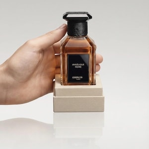 May include: A rectangular glass perfume bottle filled with amber liquid, featuring a black textured cap and a black label with the white text ANGÉLIQUE NOIRE and GGEKLAY PARIS. The bottle rests on a beige, square, textured base.