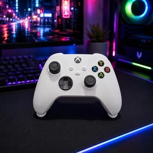 May include: A white wireless video game controller featuring a central Xbox logo button, two black analog sticks, a black directional pad, and four colored action buttons labeled Y, B, A, and X. The controller includes small menu, view, and share buttons.