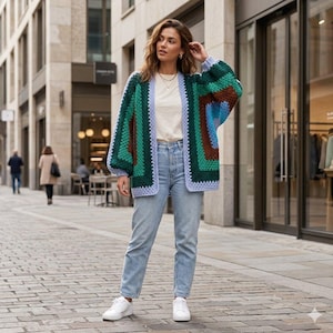 May include: A chunky crochet cardigan featuring a geometric pattern in shades of green, brown, and blue with light purple trim. The cardigan is worn over a plain white t-shirt and light blue denim jeans, paired with simple white sneakers.
