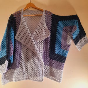May include: A crochet cardigan featuring a granny square pattern in shades of light gray, lavender, blue, and dark navy. The garment has an open front design with wide sleeves and a folded collar, constructed from interconnected yarn clusters.