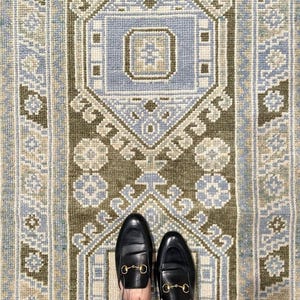 May include: A woven rug featuring a geometric pattern in olive green, light blue, and cream. The design includes a central diamond motif with nested rectangular shapes and repeating floral-like elements along the borders and vertical columns.
