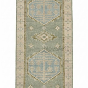 May include: A rectangular runner rug featuring a sage green field with two large light blue hexagonal medallions containing tree motifs. The border is beige with small geometric patterns. The text NovaArtIn is visible in the center.
