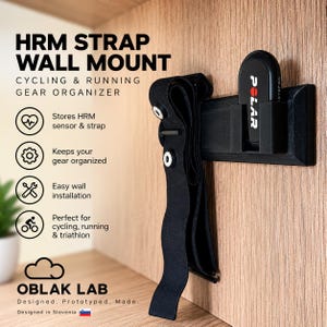 Puede incluir: Un soporte de pared negro de plástico para banda HRM con un compartimento rectangular para sensor de frecuencia cardíaca Polar y un gancho para la banda. El texto HRM STRAP WALL MOUNT CYCLING & RUNNING GEAR ORGANIZER y OBLAK LAB aparece a la izquierda.