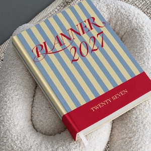 May include: A hardcover planner with a vertical light blue and cream striped pattern. The front cover features the red text "PLANNER 2027" in a script font and a solid red horizontal band at the bottom with the white text "TWENTY SEVEN".