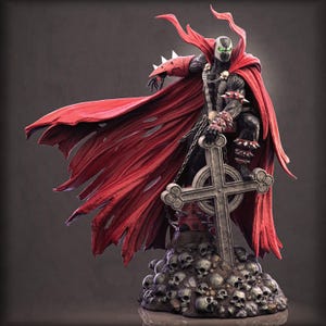 May include: A detailed Spawn action figure featuring a black textured suit with red accents, wearing a long flowing red cape. The figure stands atop a base of skulls, resting one hand on a stone cross, with spiked red gauntlets and green eyes.