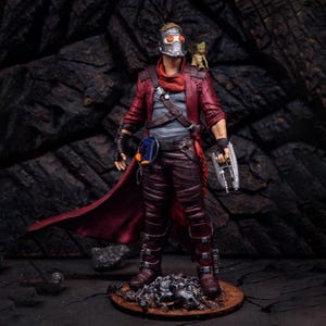 May include: A Star-Lord action figure wearing a red jacket, a silver helmet with red lenses, and a grey shirt. A small Groot figure sits on the shoulder. The figure holds a silver blaster in one hand and has a blue cassette player on the belt.