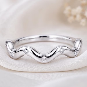 Può includere: Un anello in metallo color argento lucido caratterizzato da un design a fascia continua ondulata o serpentina. L'anello è incastonato con cinque piccole pietre rotonde trasparenti inserite a intervalli regolari lungo le creste della struttura.