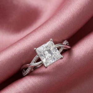 Puede incluir: Un anillo de tono plateado con una gran piedra preciosa transparente de corte cuadrado sujeta por cuatro garras. La banda tiene un diseño trenzado con pequeñas piedras transparentes engastadas en pavé, creando un aspecto brillante.