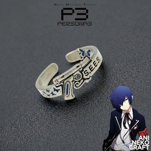 May include: A silver-toned open ring featuring a gun-shaped design with blue butterfly accents and the text S.E.E.S. engraved on the side. The ring is displayed alongside an anime-style character illustration with blue hair and a school uniform.