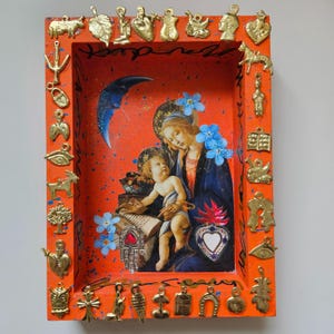 May include: An orange shadow box with a religious image of Mary and baby Jesus, decorated with gold charms. The artwork includes blue flowers and a crescent moon, with a heart-shaped emblem.