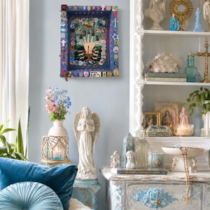May include: A rectangular shadow box wall art piece featuring a central hand illustration surrounded by small religious icons, crosses, and colorful trinkets. The frame is painted deep purple and adorned with various miniature charms and objects.
