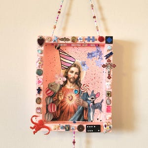 Könnte beinhalten: Ein rechteckiger, rosafarbener Schattenkastenrahmen mit einem zentralen Bild von Jesus, der einen Partyhut trägt, umgeben von verschiedenen kleinen Schmuckstücken wie einer Rehfigur, einem roten Affen, einem Kruzifix und bonbonartigen Dekorationen.