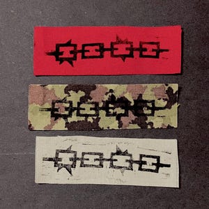 May include: Three rectangular fabric patches featuring a black printed graphic of a horizontal chain with four square links and sharp spikes. The top patch is solid red, the middle patch has a camouflage pattern, and the bottom patch is off-white.