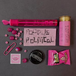 May include: A collection of items including a pink bubble wand, a pink shotgun shell labeled Top Gun 2 3/4 1 1/8-8, a pink fabric patch with the text Food Is Political, pink beads, a pink paperclip, a square sticker, a black bottle cap, and a candy wrapper.