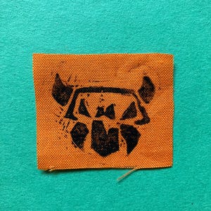 May include: A rectangular orange fabric patch featuring a black ink-stamped graphic of a stylized, geometric creature head with pointed ears and angular facial features. The woven texture of the orange textile is clearly visible throughout the patch.
