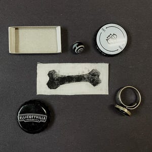 May include: A collection of small items including an empty rectangular cardboard box, a black and white swirled marble, a white bottle cap with black text, a rectangular fabric patch with a black bone print, a black Ellicottville Brewing Co. bottle cap, and a metal ring.