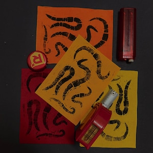May include: Three rectangular fabric swatches in orange, yellow, and red, each featuring a black block-printed worm pattern. A red bottle cap with a white R, a red lighter, and a red measuring tape showing metric units are placed on top.