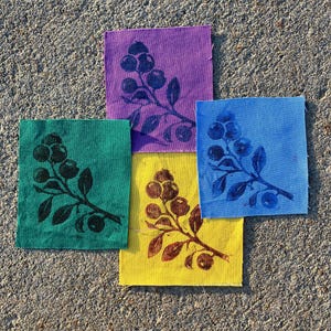May include: Four square fabric patches featuring a botanical print of a berry branch. The patches are colored in purple, green, yellow, and light blue, each with a contrasting ink print of the same berry branch design in dark tones.
