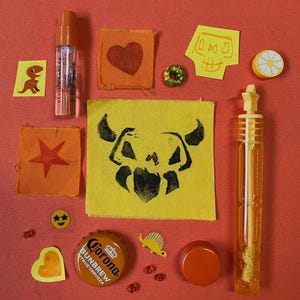 May include: A collection of small items including a clear spray bottle, orange and yellow fabric patches with heart, star, and skull prints, a Corona Sunbrew bottle cap, a yellow bubble wand with a star topper, and various small stickers and beads.