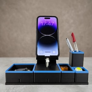 Può includere: Un organizer da scrivania nero con bordi blu brillante, dotato di un supporto centrale per smartphone con cavo di ricarica, un portapenne e tre scomparti aggiuntivi per cancelleria, con uno smartphone che mostra l'orario 17:47.