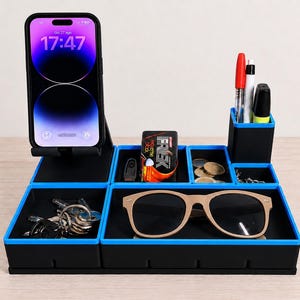 Può includere: Un organizer da scrivania nero con bordi blu brillante, dotato di supporto verticale per smartphone, portapenne alto e diversi scomparti contenenti chiavi, occhiali da sole, monete e una scatola con etichetta Frik Extreme Black.