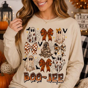 Op de afbeelding: Een beige sweatshirt met lange mouwen en een grafische print van vier spoken, drie strikken met patronen, spinnenwebben en kleine vleermuizen. De tekst "BOO-JEE" is onderaan het ontwerp gedrukt in een bruin, druipend lettertype.
