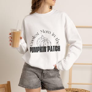 Peut inclure: Un sweat-shirt blanc à manches longues avec une impression graphique noire représentant deux citrouilles et le texte Coolest Mom in the PUMPKIN PATCH. Le vêtement possède un col rond et des manches longues à bords-côtes, porté avec un short en jean gris.