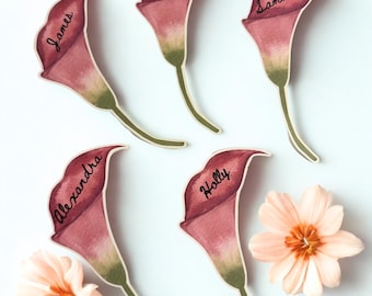 Op de afbeelding: Vijf gestanste papieren naamkaartjes in de vorm van calla lelies in tinten donkerroze en groen. Elke lelie is voorzien van een handgeschreven naam in zwarte inkt: James, Mike, Samantha, Alexandra en Holly. Twee licht perzikkleurige bloemen liggen ernaast.