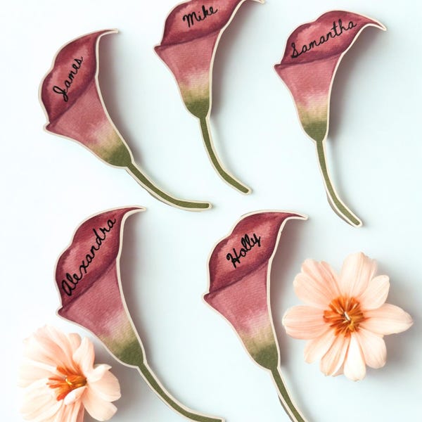 May include: Five die-cut paper place cards shaped like calla lilies in shades of deep pink and green. Each lily features a handwritten name in black ink: James, Mike, Samantha, Alexandra, and Holly. Two pale peach flowers are placed nearby.