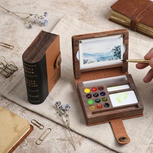 May include: A portable wooden watercolour set shaped like a small book with a leather cover. The set includes a palette with twelve small paint wells, two mixing areas, and a small landscape painting clipped to the top. The spine reads MINI BOOK.