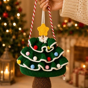 Op de afbeelding: Een gehaakte tas met trekkoord in de vorm van een kerstboom met een donkergroen gelaagd lichaam, witte rand en kleurrijke pompons. Het heeft een gele ster aan de bovenkant en een bruine basis, opgehangen aan twee rood-wit gedraaide touwhengsels.