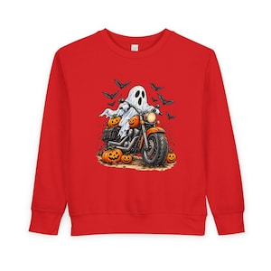 Puede incluir: Una sudadera roja de manga larga con un estampado gráfico de un fantasma blanco montado en una motocicleta. La escena incluye varias calabazas naranjas talladas en el suelo y una bandada de murciélagos negros volando en el fondo.