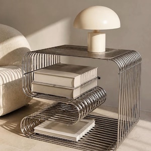 Pode incluir: Uma mesa lateral de metal prateado com um design de estrutura de arame curvo em forma de S. A mesa possui várias superfícies horizontais que sustentam três livros brancos. Um candeeiro de mesa em forma de cogumelo cor de creme está no topo.