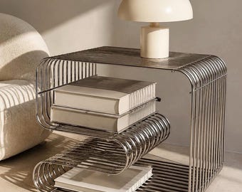 Può includere: Un tavolino in metallo color argento con una struttura in filo metallico curva a forma di S. Il tavolo presenta diverse superfici orizzontali che ospitano tre libri bianchi. Una lampada da tavolo a forma di fungo color crema è posizionata sopra.